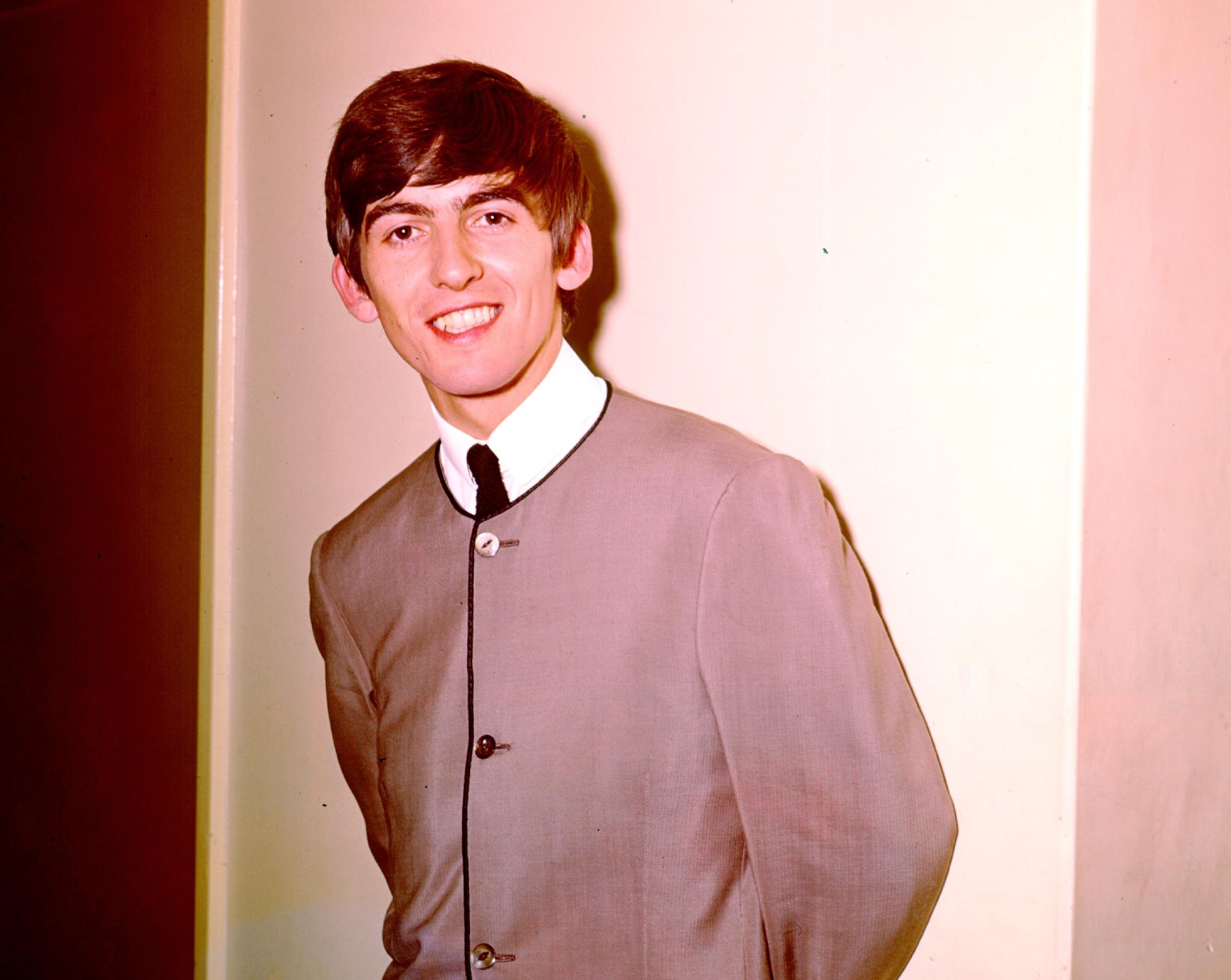 George Harrison wears a jacket and tie and stands in front of a white wall.