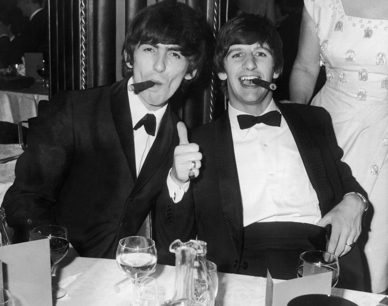 A black and white picture of George Harrison and Ringo Starr smoking cigars at a dinner table.