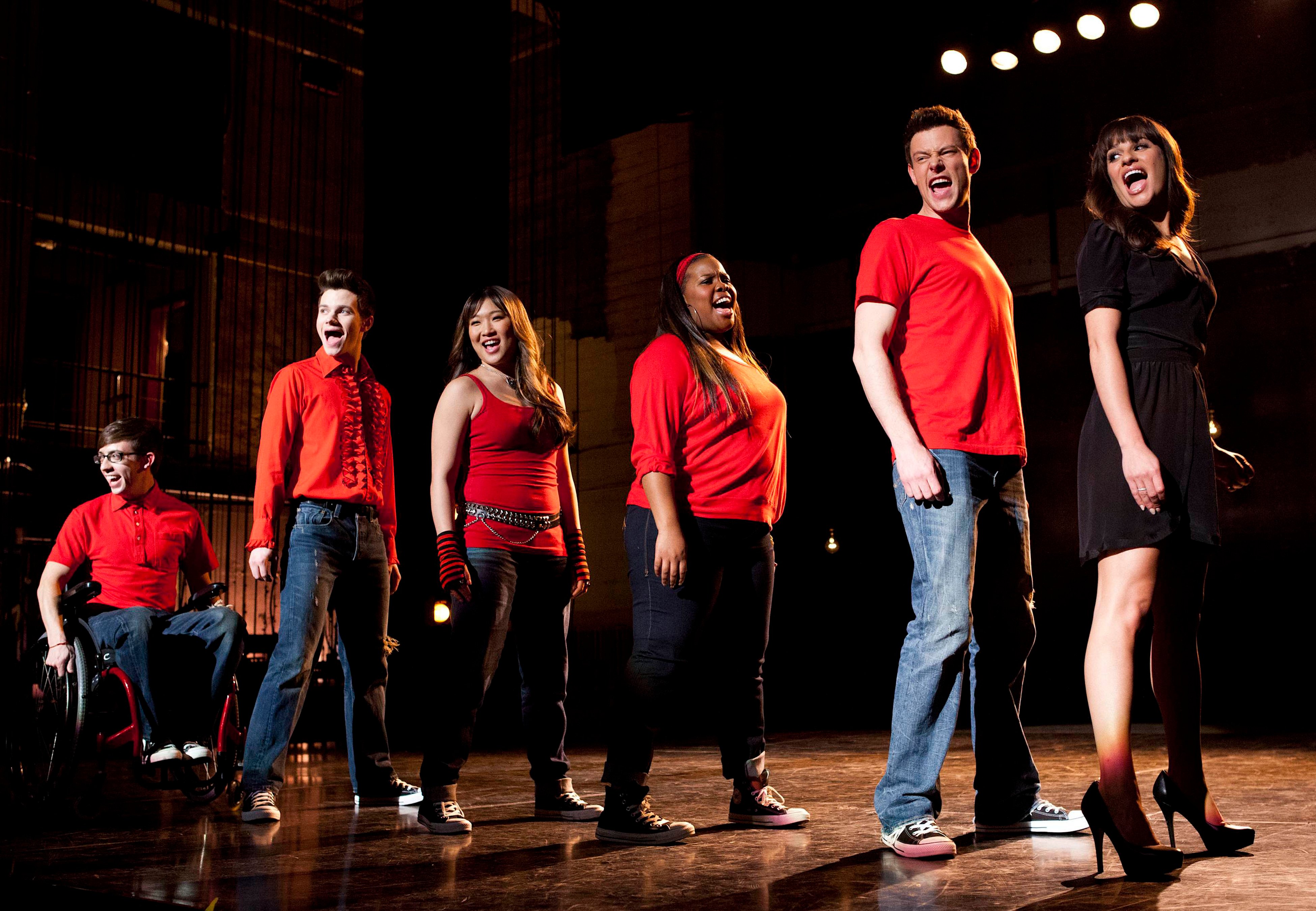 Kevin McHale, Chris Colfer, Jenna Ushkowitz, Amber Riley, Cory Monteith, and Lea Michele, in character as Artie, Kurt, Tina, Mercedes, Finn, and Rachel, share a scene in 'Glee' Season 4, which is now on Hulu and Disney+. The characters in the photo stand in a line onstage and sing.