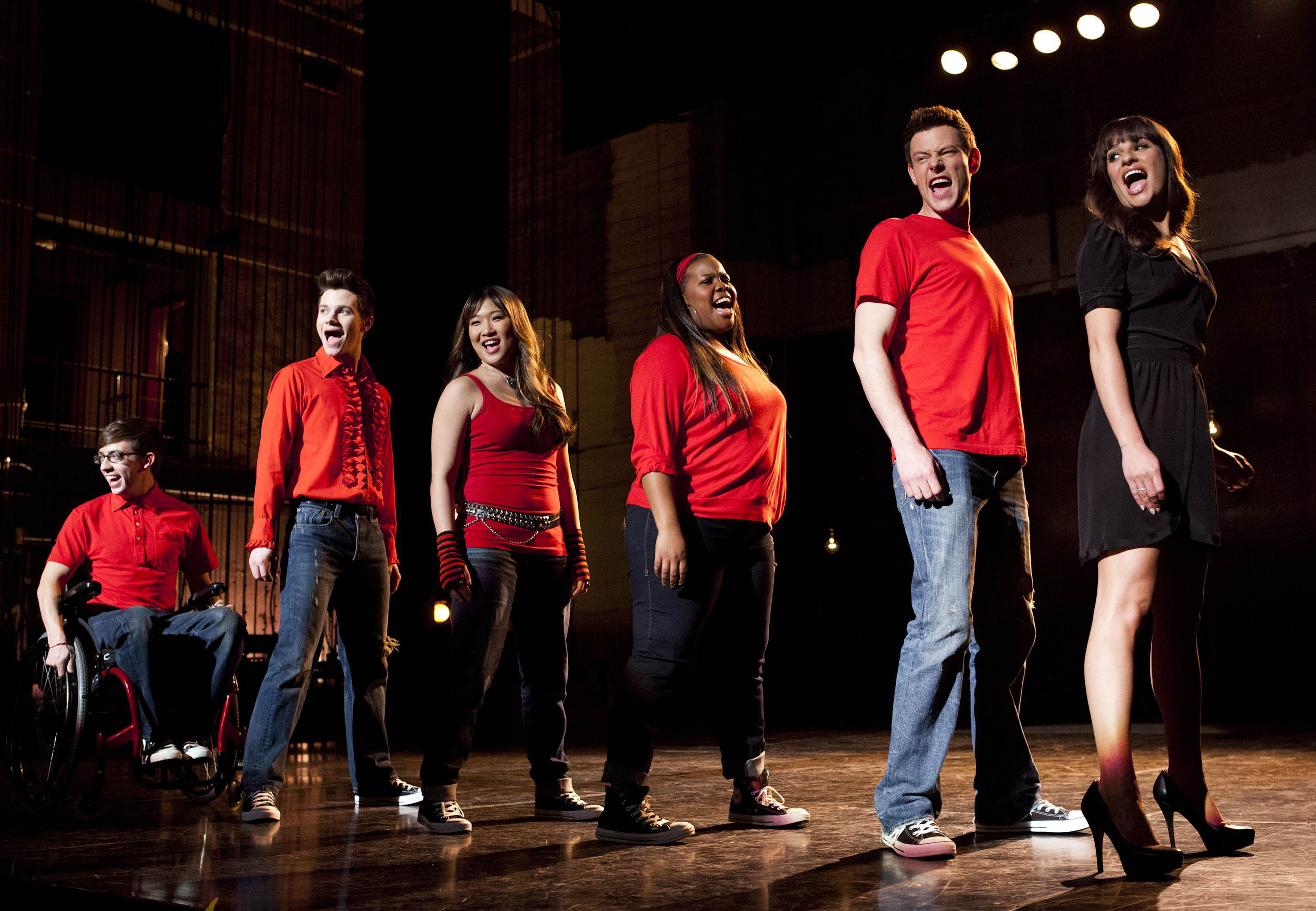 Kevin McHale, Chris Colfer, Jenna Ushkowitz, Amber Riley, Cory Monteith, and Lea Michele, in character as Artie, Kurt, Tina, Mercedes, Finn, and Rachel, share a scene in 'Glee' Season 4, which is now on Hulu and Disney+. The characters in the photo stand in a line onstage and sing.