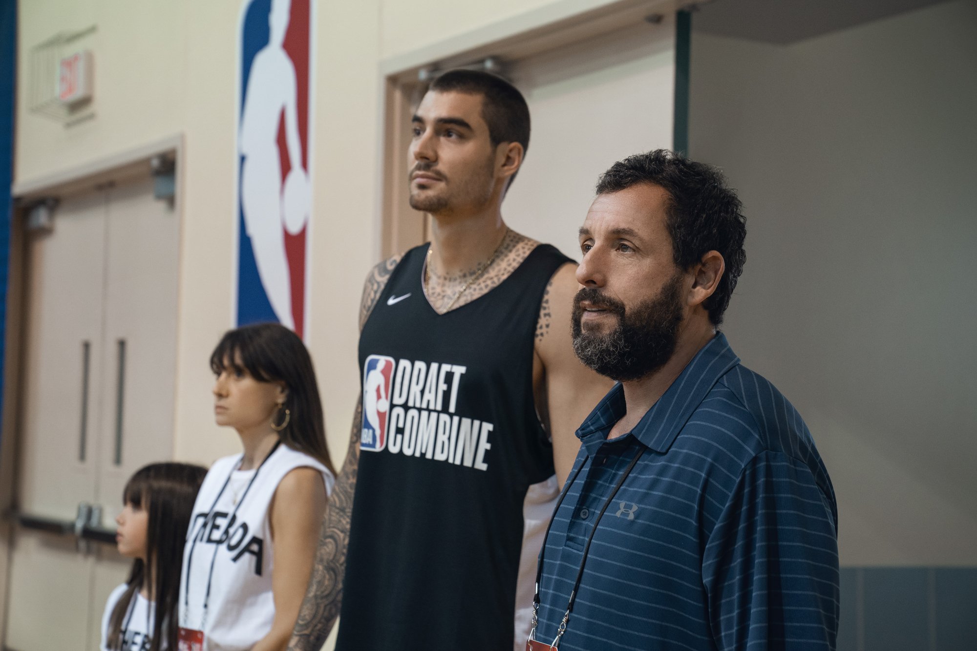 'Hustle' Ainhoa Pillet as Lucia, Maria Botto as Paola, Juancho Hernangomez as Bo Cruz and Adam Sandler standing in front of the NBA logo looking out at the court.