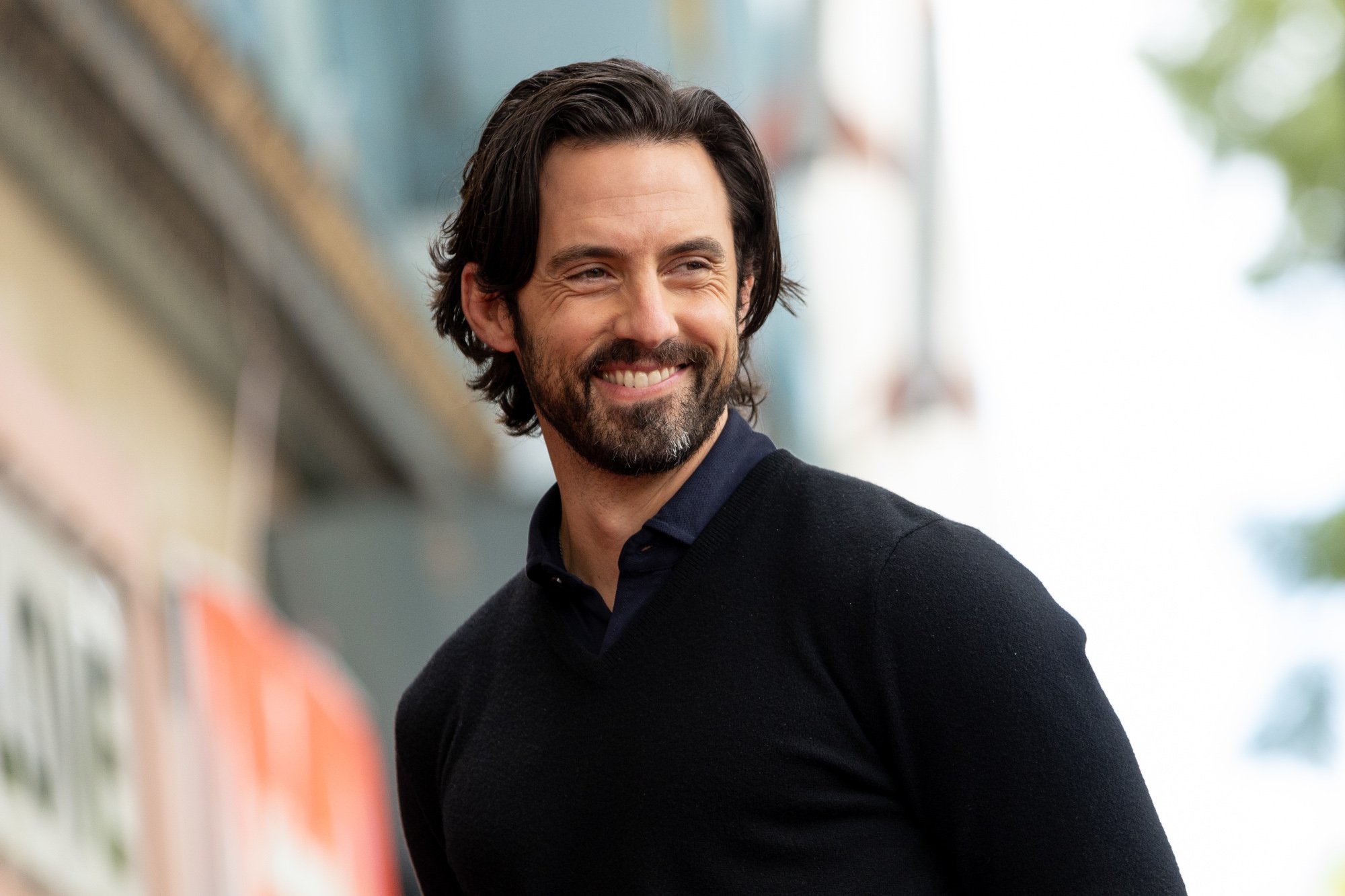 'This Is Us' star Milo Ventimiglia, who plays Jack Pearson on the show. He's wearing a black shirt, looking to the side of the camera, and smiling.