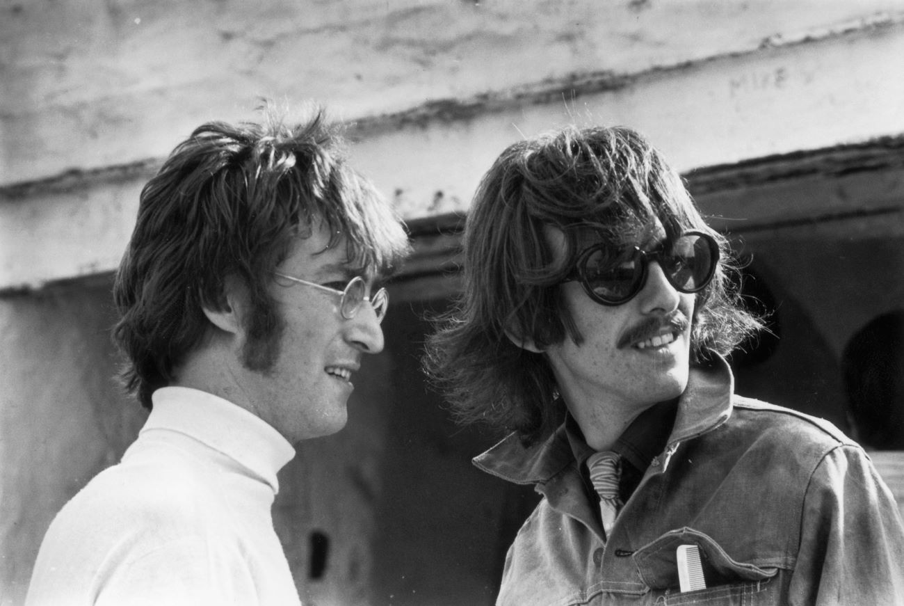 A black and white picture of John Lennon and George Harrison. Lennon wears a turtleneck and glasses and Harrison wears a jacket and sunglasses.