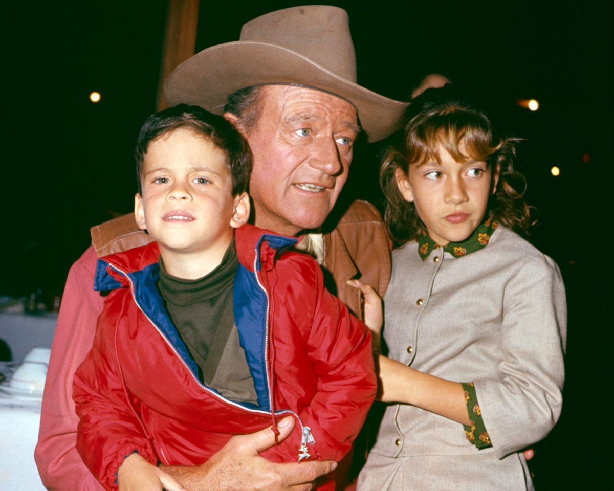 John Wayne, Ethan Wayne, and Aissa Wayne. Aissa wrote about her father's masculinity. John is wearing a cowboy hat and holding Ethan and Aissa in each arm.