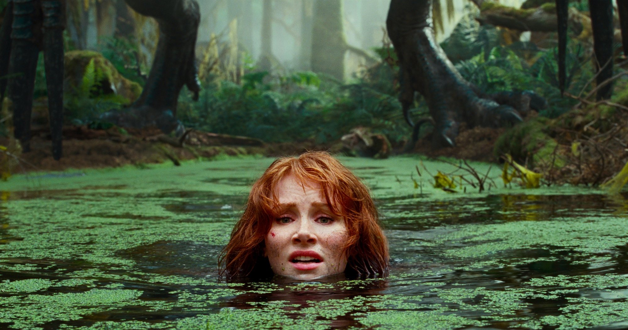 'Jurassic World Dominion' Bryce Dallas Howard as Claire Dearing with her head above water with moss on the surface. A pair of dinosaur legs are standing behind her on the land.