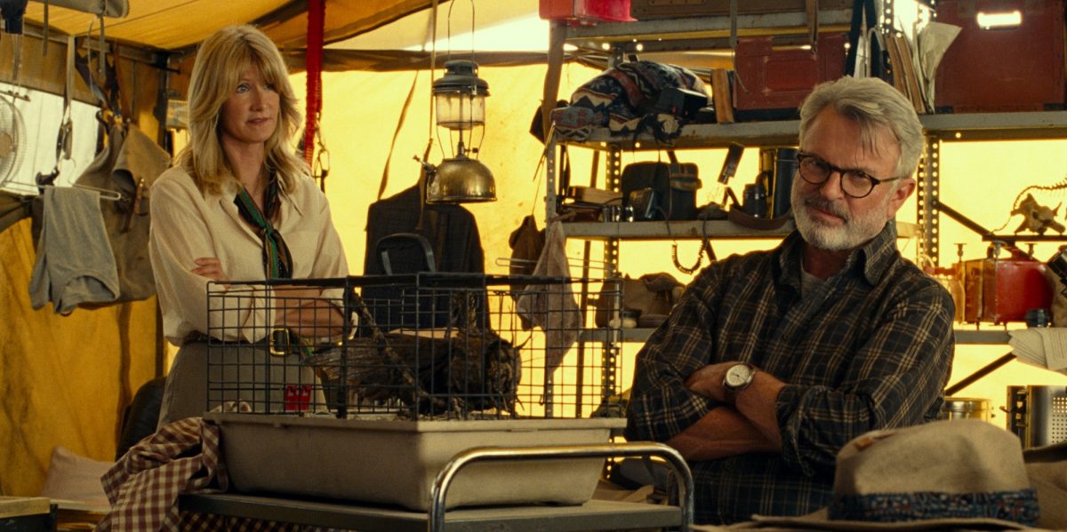 'Jurassic World Dominion' Laura Dern as Dr. Ellie Sattler and Sam Neill as Dr. Alan Grant in a tent surrounded by dig site items with their arms crossed.