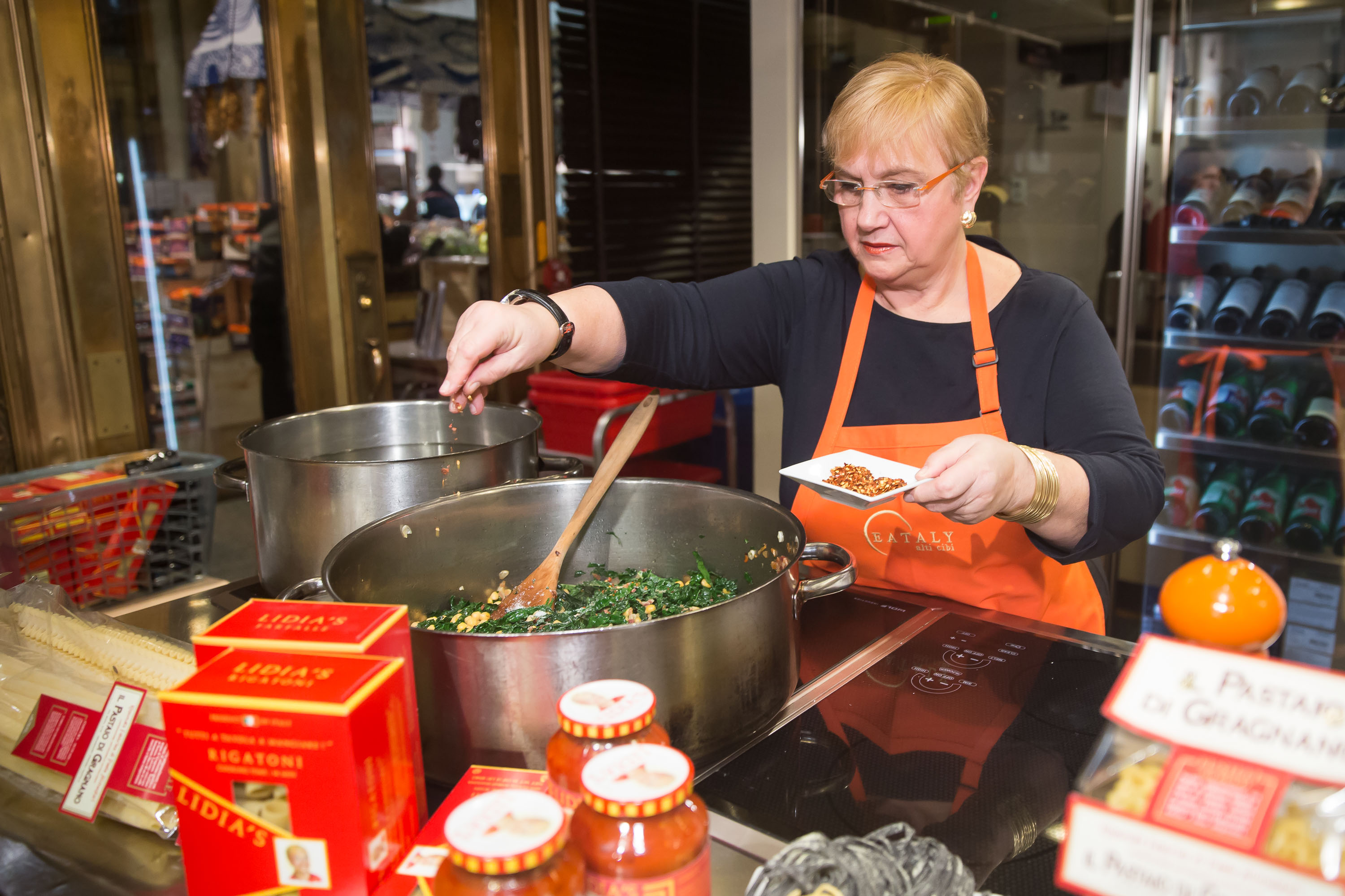 Celebrity chef Lidia Bastianich wears an orange apron as she prepares a dish in this photograph.