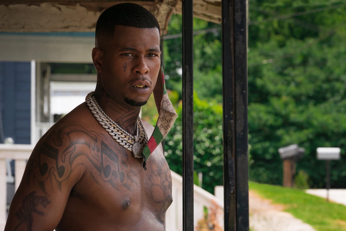 J. Alphonse Nicholson appears in a scene from 'P-Valley' Season 2 as his character Lil Murda. He's standing outside on a porch wearing a Gucci face mask.