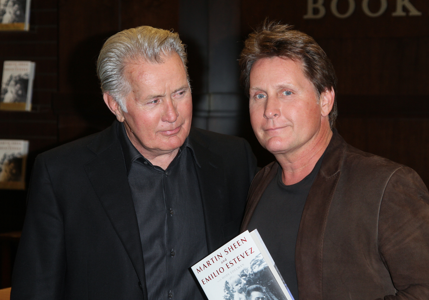 Martin Sheen (L) and Emilio Estevez (R) smiling together and holding a book