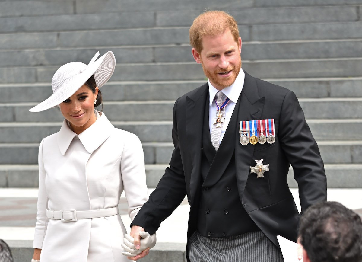 Meghan Markle wears a white coat and hat while smiling and holding hands with Prince Harry dressed in a dark suit and tie
