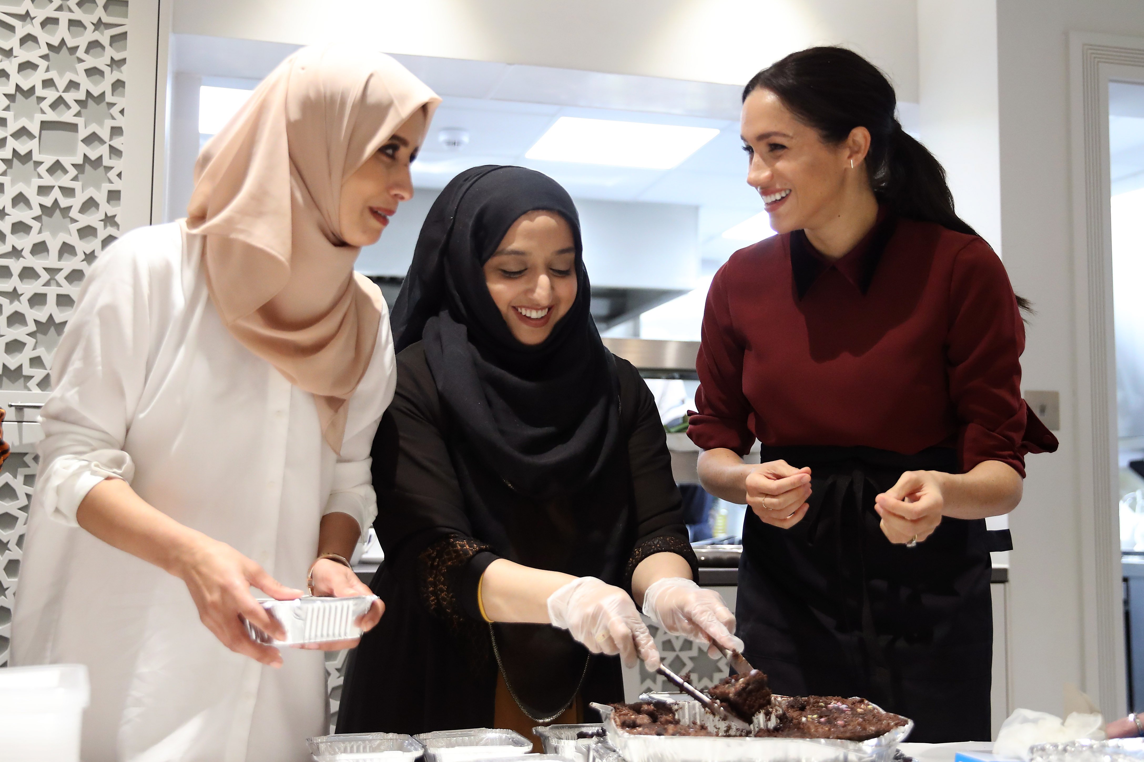 Meghan Markle (R) at the charity organization Hubb Community Kitchen smiling and preparing food while wearing a red sweater.