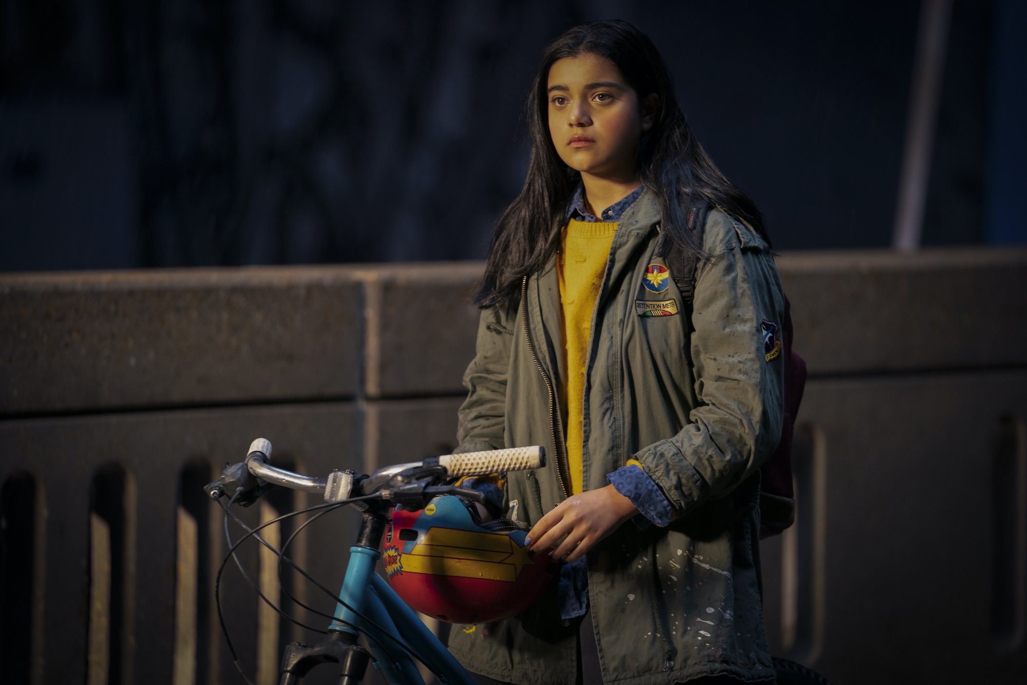 Iman Vellani, in character as Kamala Khan in 'Ms. Marvel' on Disney+, wears a gray jacket over a yellow sweater while sitting on her bike.