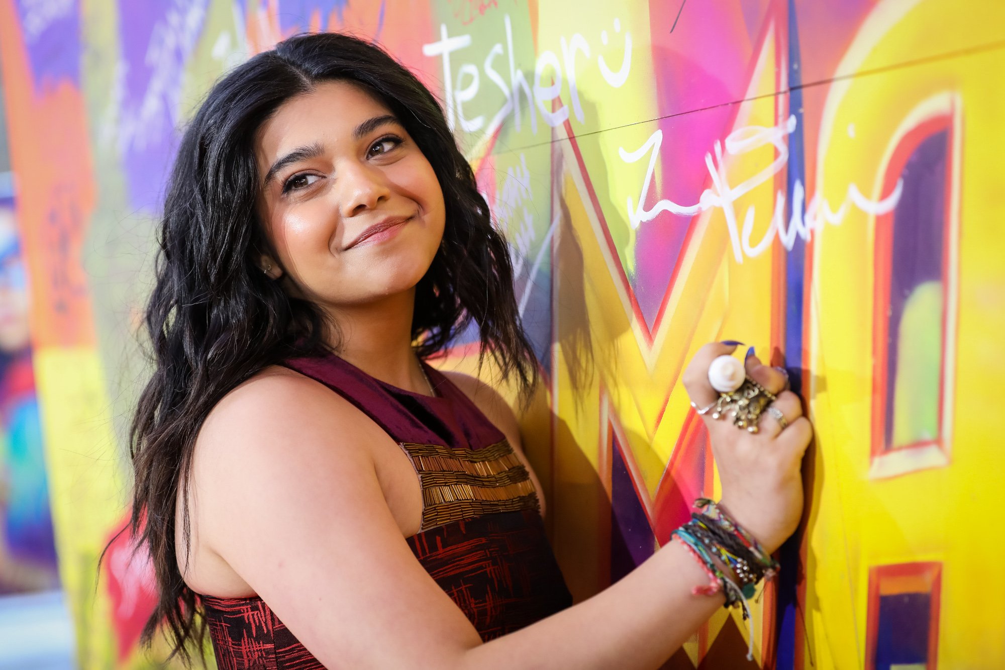 'Ms. Marvel' star Iman Vellani at the Disney+ show's screening. She's wearing a red and black dress with gold on the front, and she's leaning against a brightly colored wall.