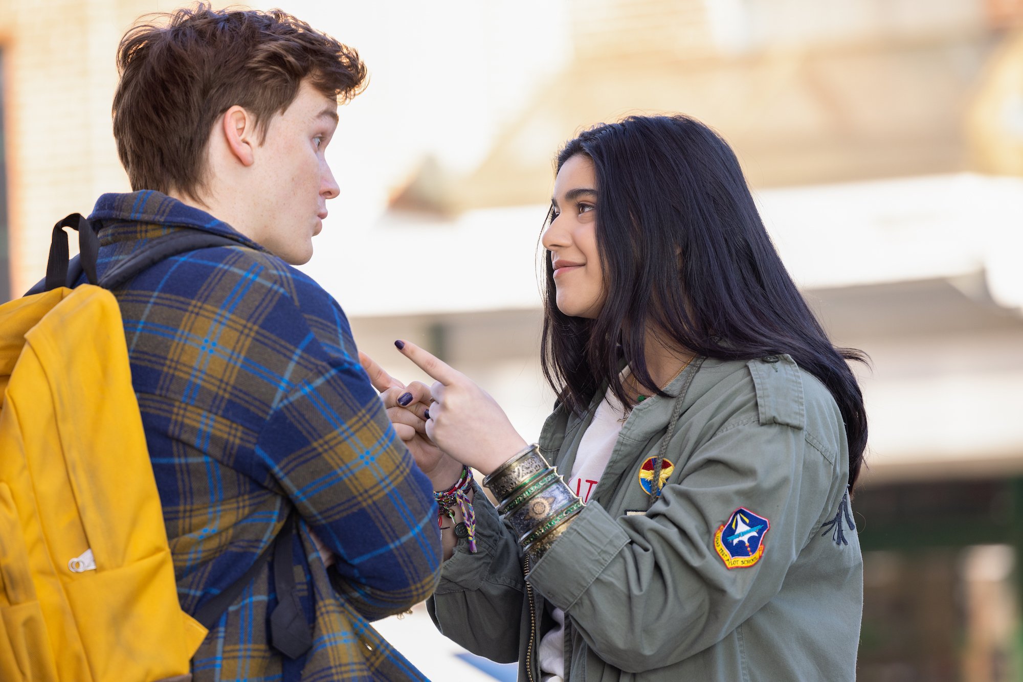 Matthew Lintz and Iman Vellani, in character as Bruno and Kamala, share a scene in 'Ms. Marvel' Episode 2. Bruno wears a blue and yellow plaid shirt and yellow backpack. Kamala wears an army green jacket over a white shirt.
