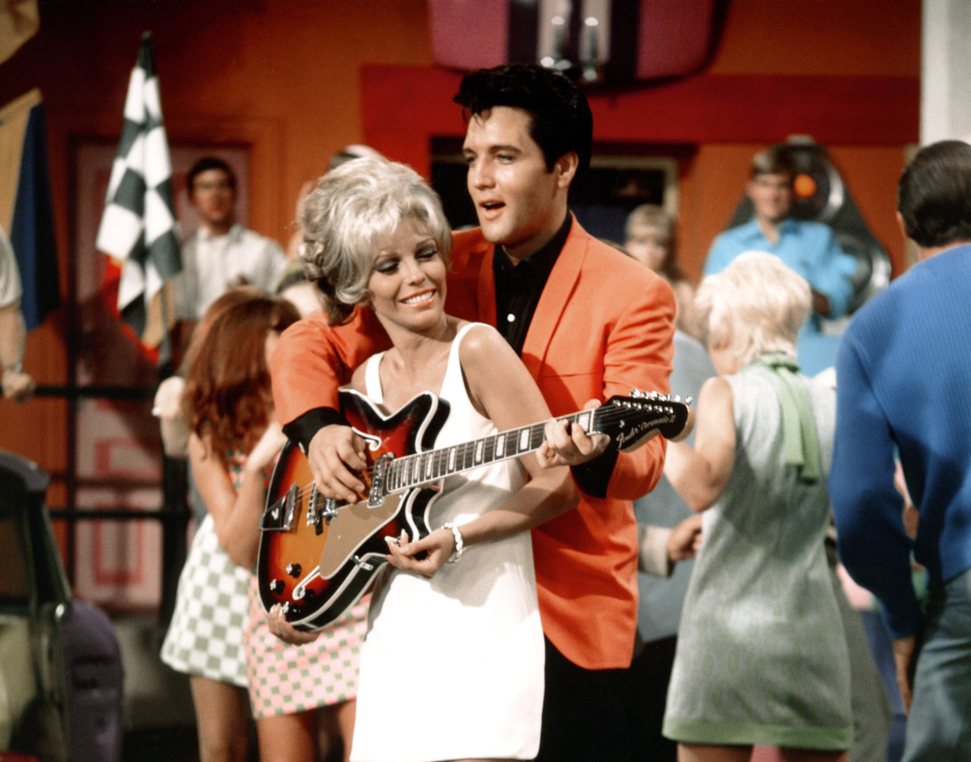 Nancy Sinatra wears a white dress and stands in front of Elvis, who holds his guitar in front of her.
