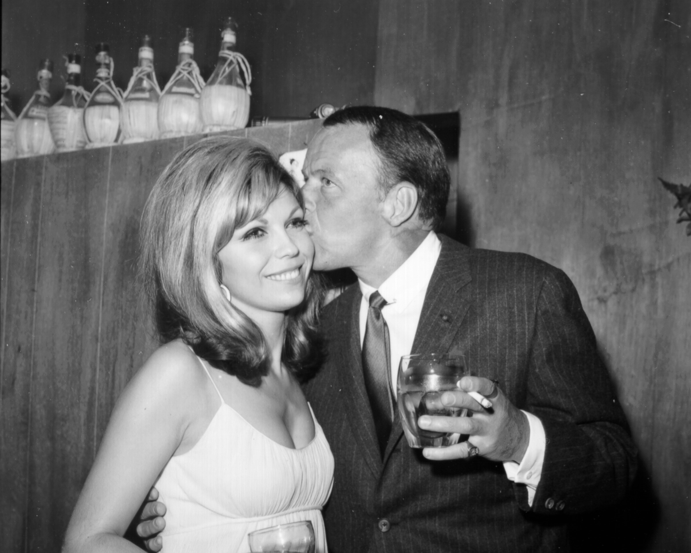 A black and white picture of Frank Sinatra holding a drink and a cigarette and kissing Nancy Sinatra on the cheek.