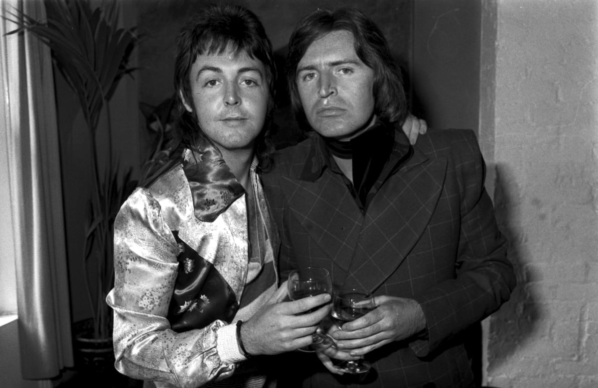 Paul McCartney and his brother Mike hold wine glasses and stand next to eachother while smiling.