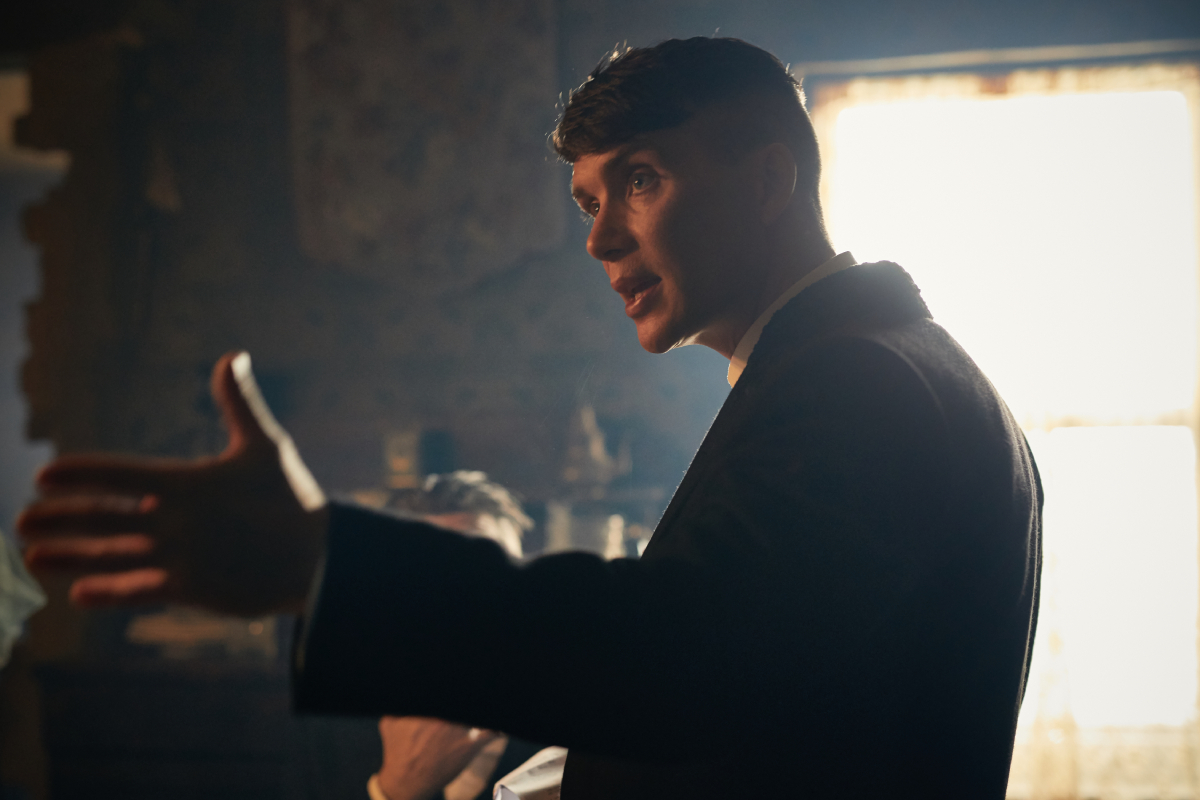 Understanding the timeline is important in Peaky Blinders. Tommy Shelby stands in a house with a hand raised. 