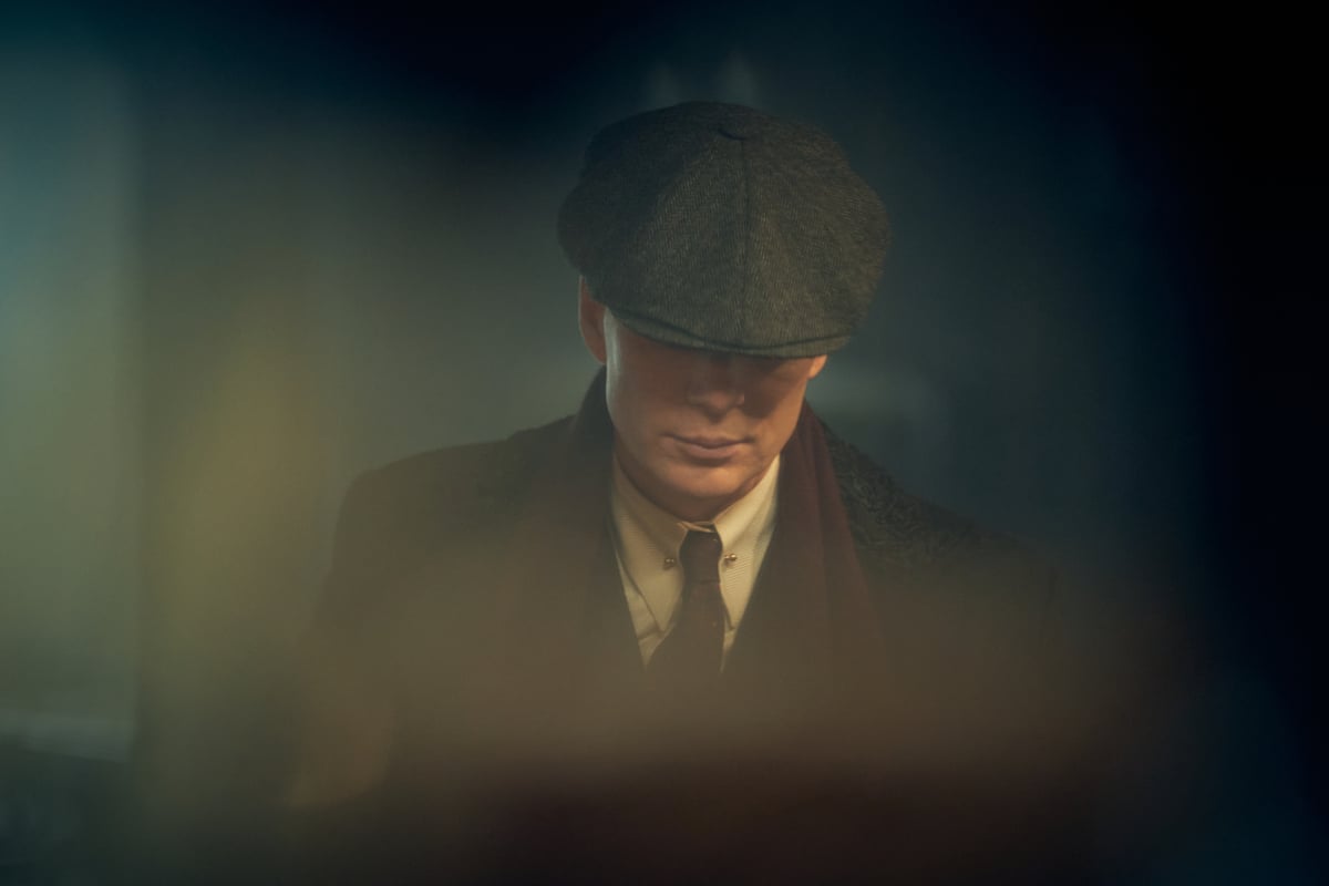 Cillian Murphy as Tommy Shelby in Peaky Blinders Season 6. Tommy wears a suit, red scarf, and flat-billed cap. 