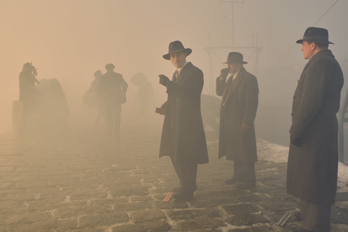 Michael Gray smoking a cigarette in Peaky Blinders Season 6. Michael stands near a boat with two men behind him.