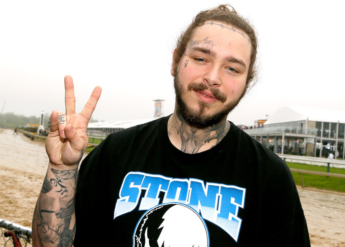 Post Malone smiling and holding a peace sign with his fingers while standing outside. Post Malone and his fiancée are getting a lot of attention since he announced the birth of his baby girl in 2022.