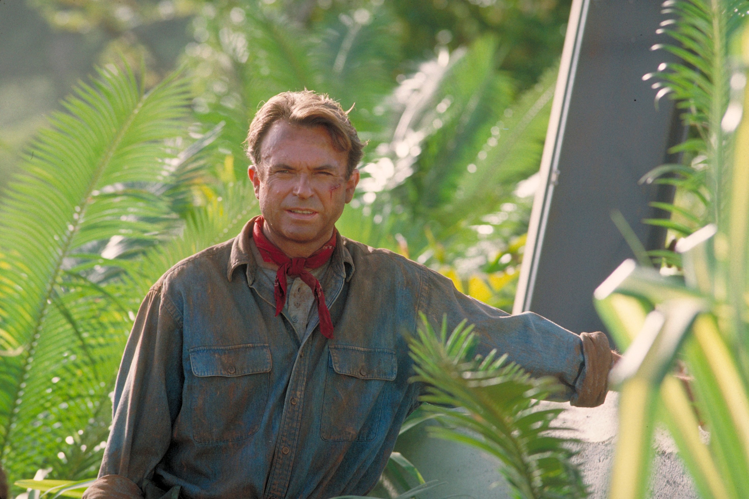 Jurassic World Dominion actor Sam Neill stands next to an electric fence as Dr. Alan Grant in Jurassic Park