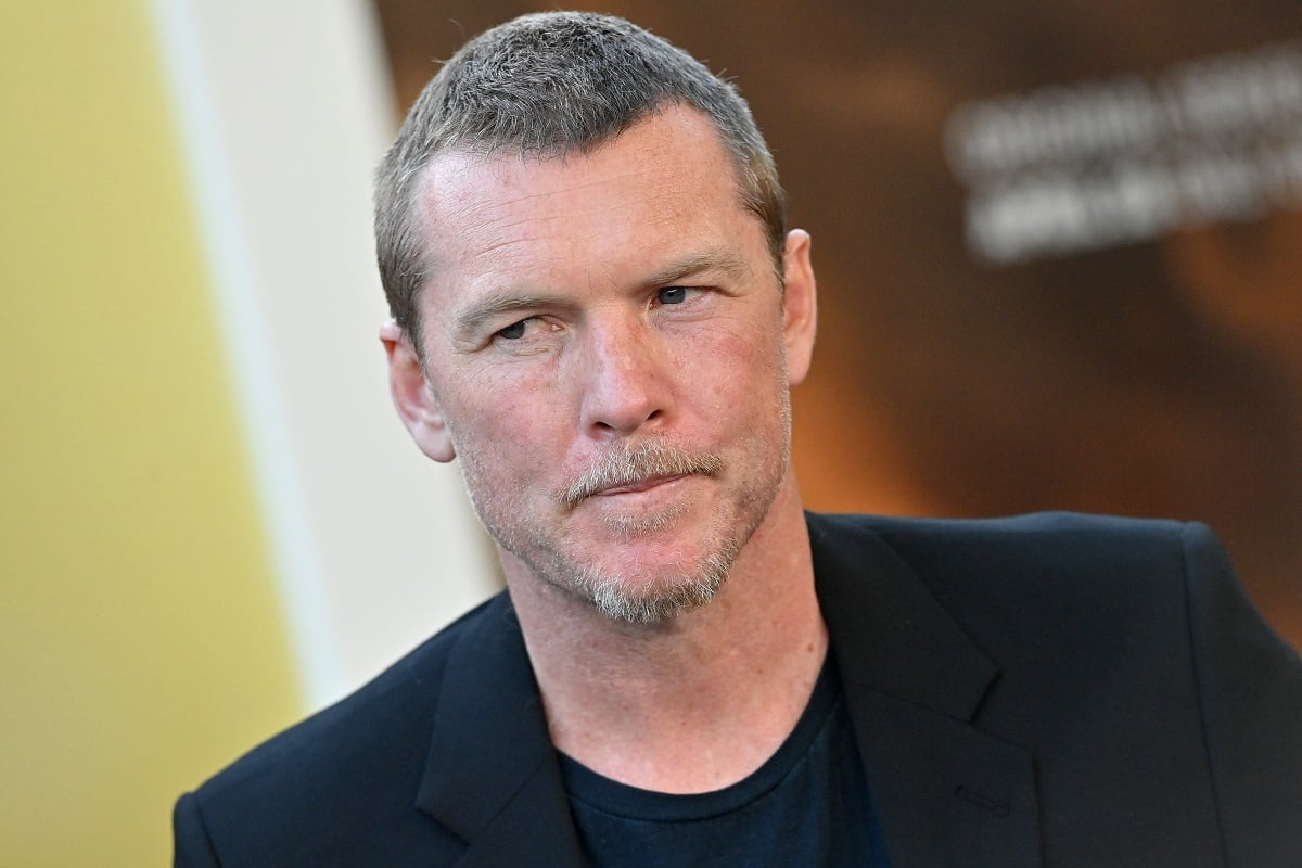 Sam Worthington posing while wearing a black shirt and blazer.