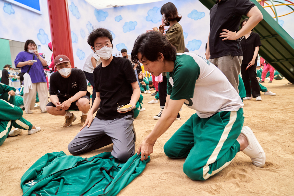 The 'Squid Game' cast and crew filming a game with Lee Jung-jae kneeling in sand and placing his green jacket down