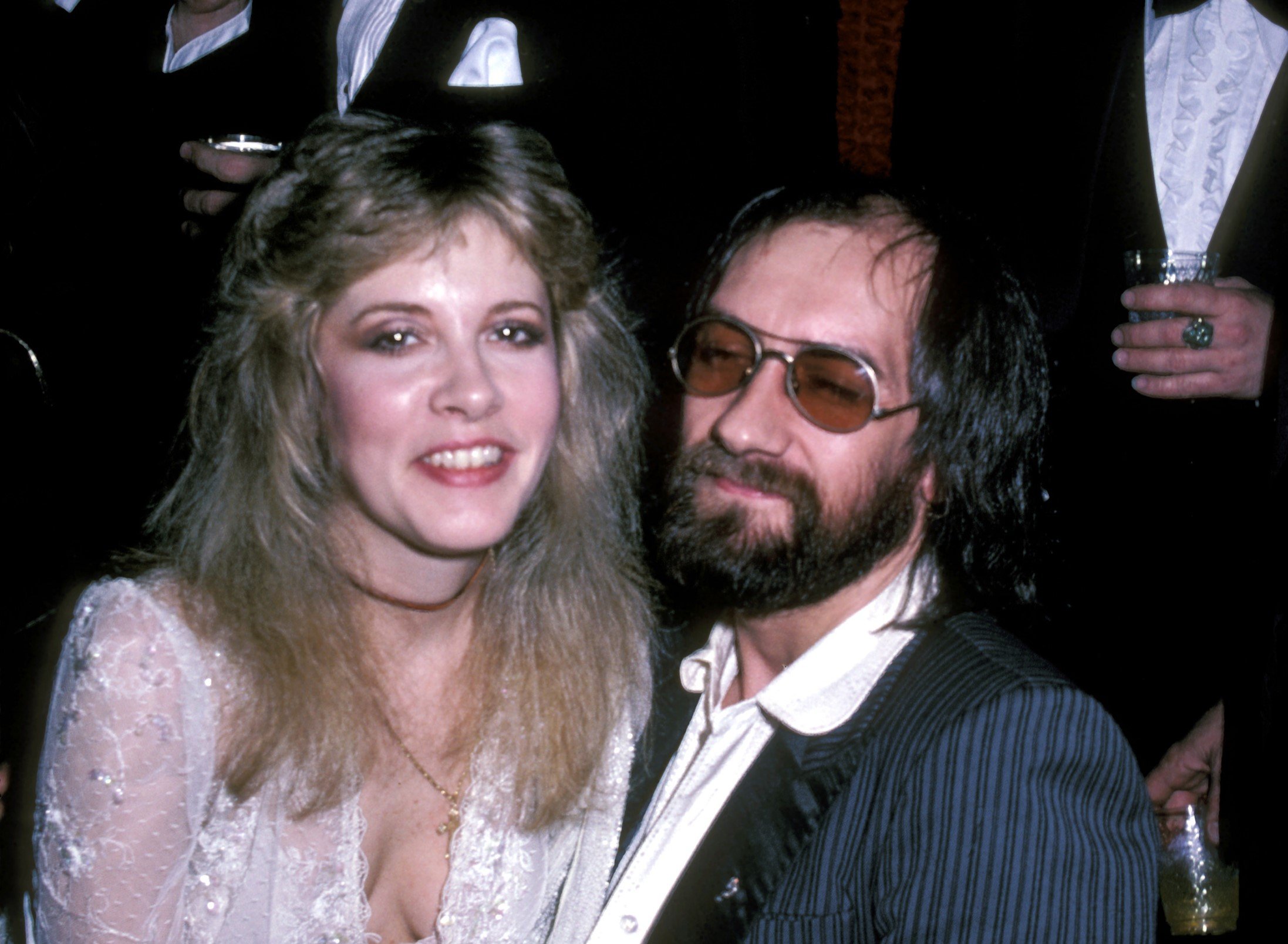 Stevie Nicks wears a white lace dress and sits on Mick Fleetwood's lap. He wears sunglasses.