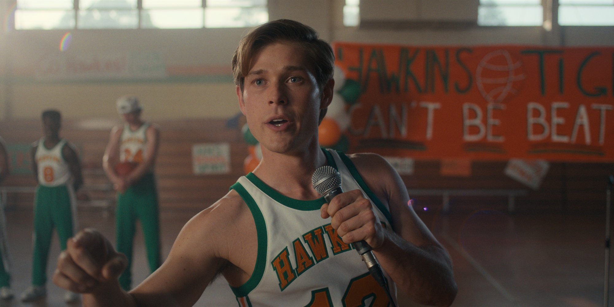 'Stranger Things 4' star Mason Dye wearing a basketball uniform and holding a microphone in a production still.