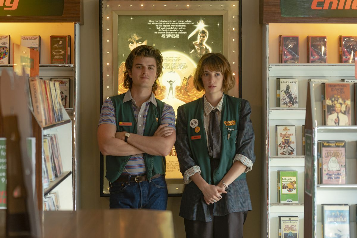 'Stranger Things' Robin Buckley and Steve Harrington stand in front of a movie poster while wearing their Family Video uniforms in a production still from season 4.