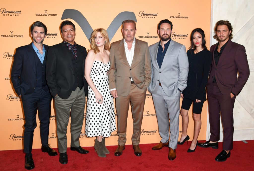 The Yellowstone cast stands in front of the Yellowstone logo at a red carpet event.