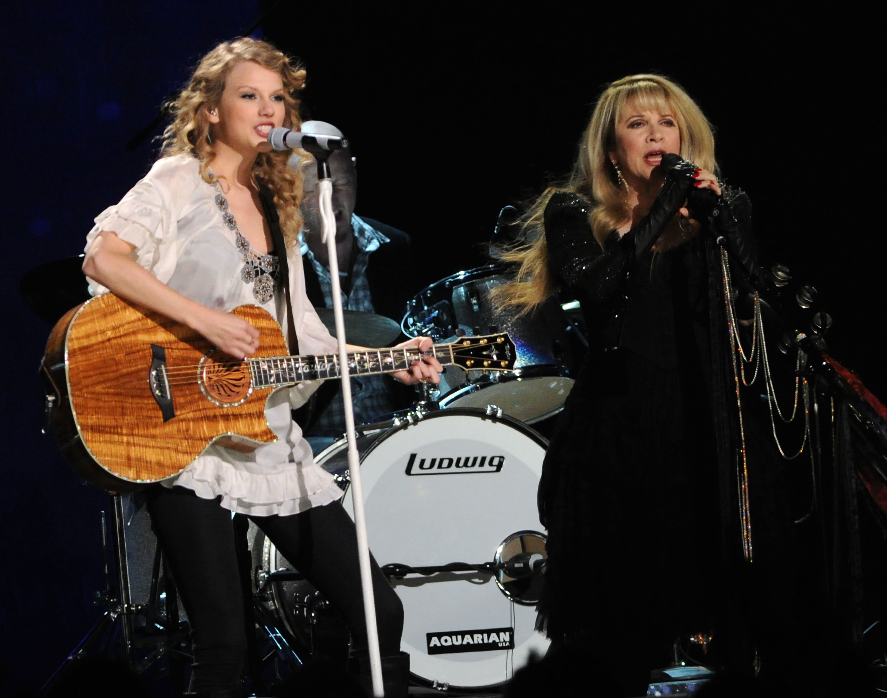 Taylor Swift wears a white shirt and strums a guitar in front of a microphone. Stevie Nicks wears a black dress and sings into a microphone.