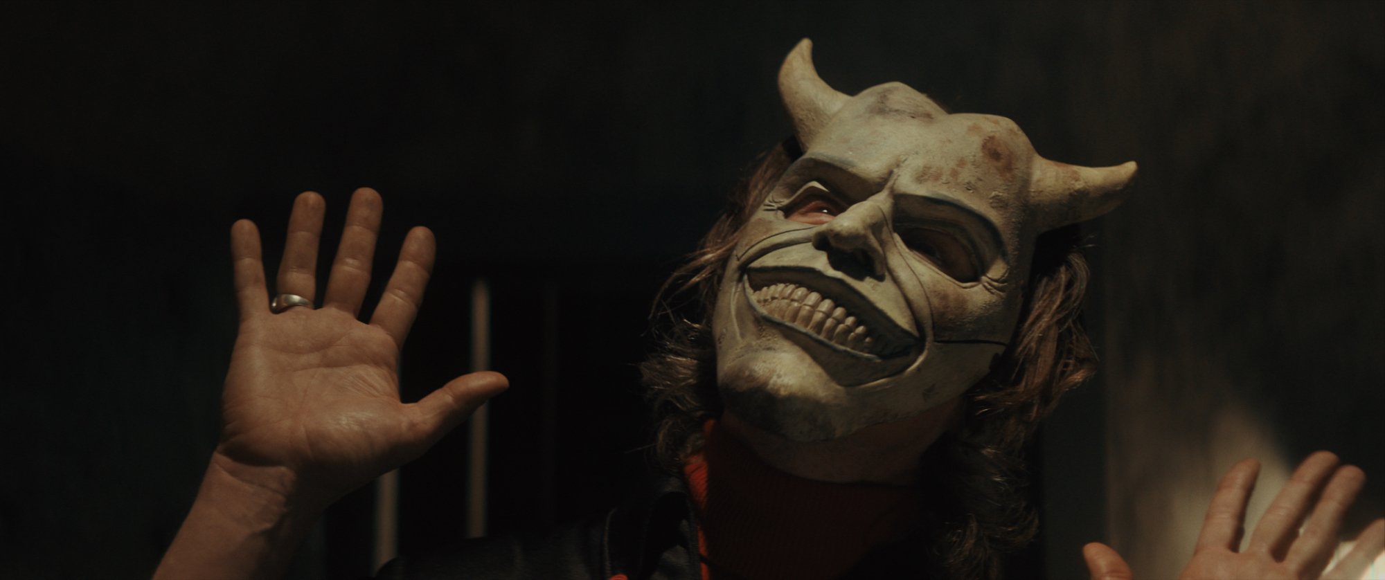Ethan Hawke as The Grabber in The Black Phone, directed by Scott Derrickson. looking up wearing a demon mask with his open hands held up