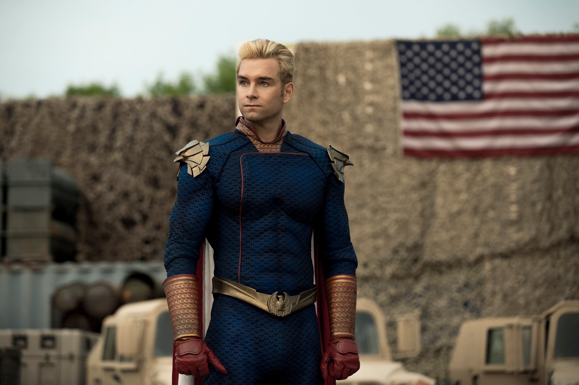Antony Starr as Homelander in 'The Boys' Season 2. He's wearing a blue suit adorned with red and gold, and there's an American flag in the background.