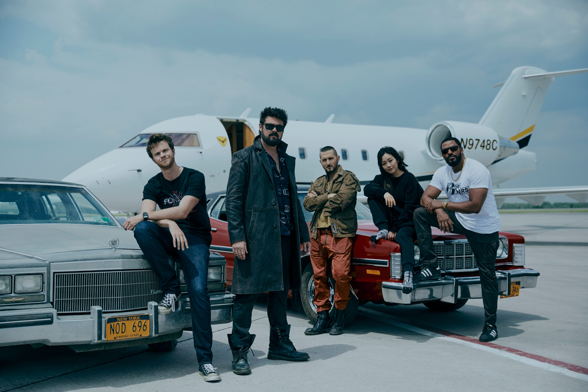 Jack Quaid (Hughie Campbell), Karl Urban (Billy Butcher), Tomer Capone (Frenchie), Karen Fukuhara (Kimiko), Laz Alonso (Mother's Milk) standing in front of an airplane in an image for 'The Boys,' which was renewed for season 4.