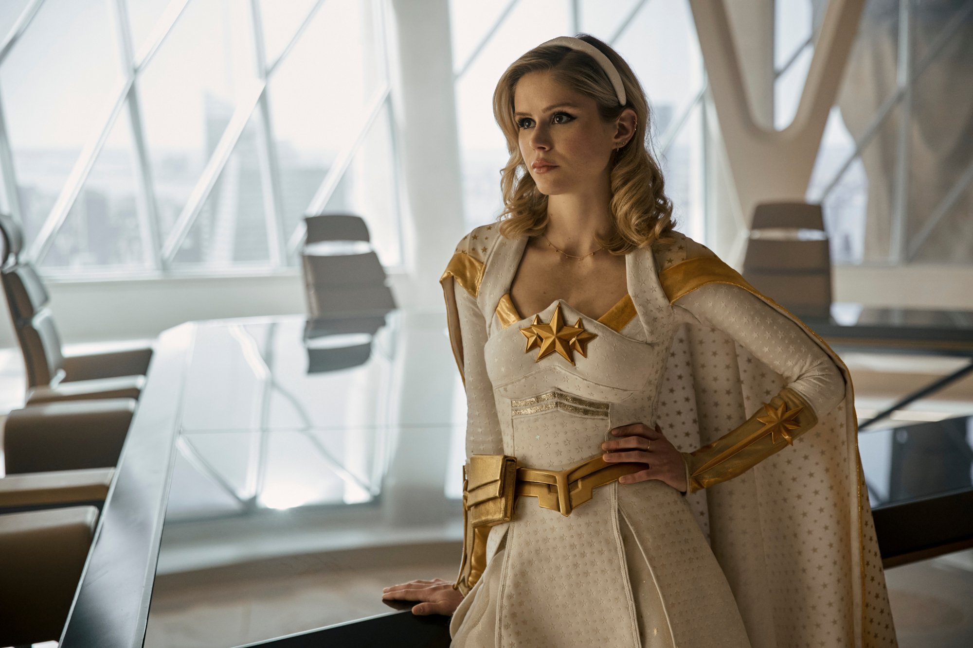 Erin Moriarty in 'The Boys' Season 3, which just streamed its Herogasm episode. She's wearing her white and gold costume and leaning against a table with her hand on her hip.