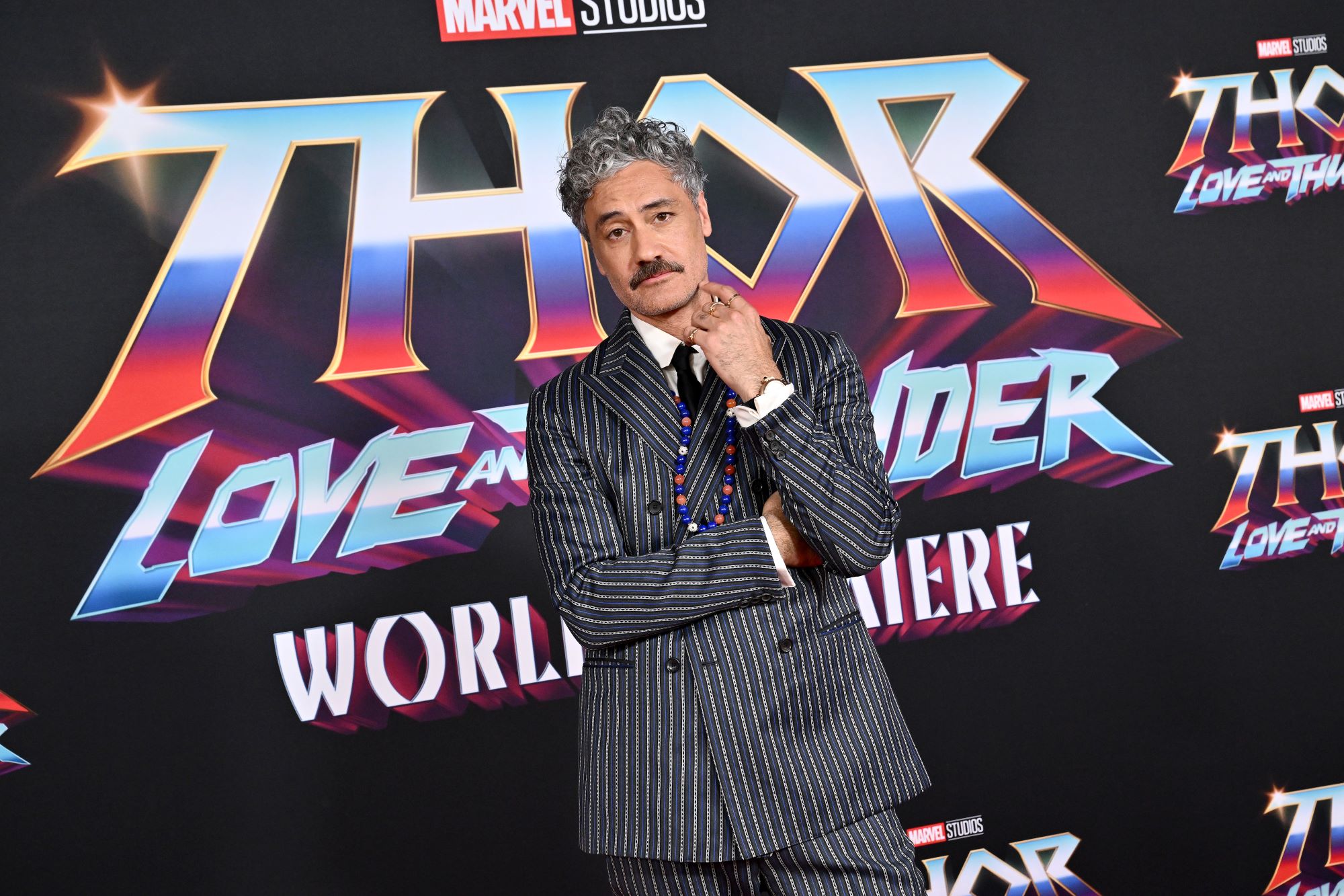 Taika Waititi, who says there are major cameos in 'Thor: Love and Thunder,' wears a dark gray striped suit over a white button-up shirt, black tie, and blue, orange, and white beads.