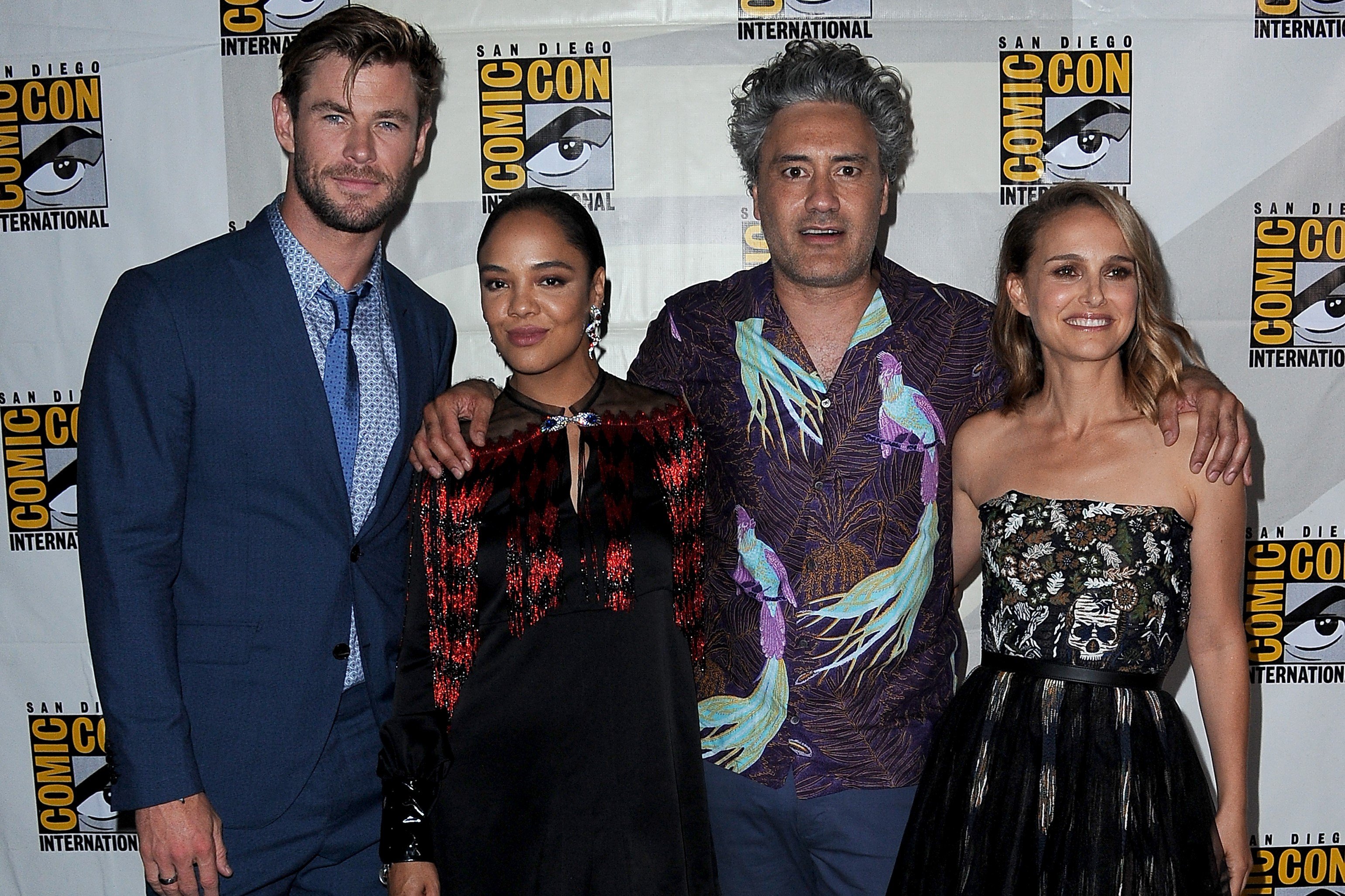Chris Hemsworth, Tessa Thompson, Taika Waititi, and Natalie Portman, the cast of 'Thor: Love and Thunder,' which includes the Guardians of the Galaxy, pose for pictures. Hemsworth wears a blue suit. Thompson wears a black and red long-sleeved dress. Waititi wears a blue and purple bird patterned shirt. Portman wears a black and white strapless dress.