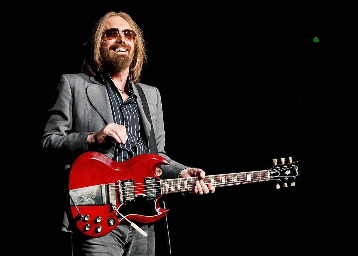 Tom Petty stands on stage wearing sunglasses and holding a red guitar, preparing to play music in concert.