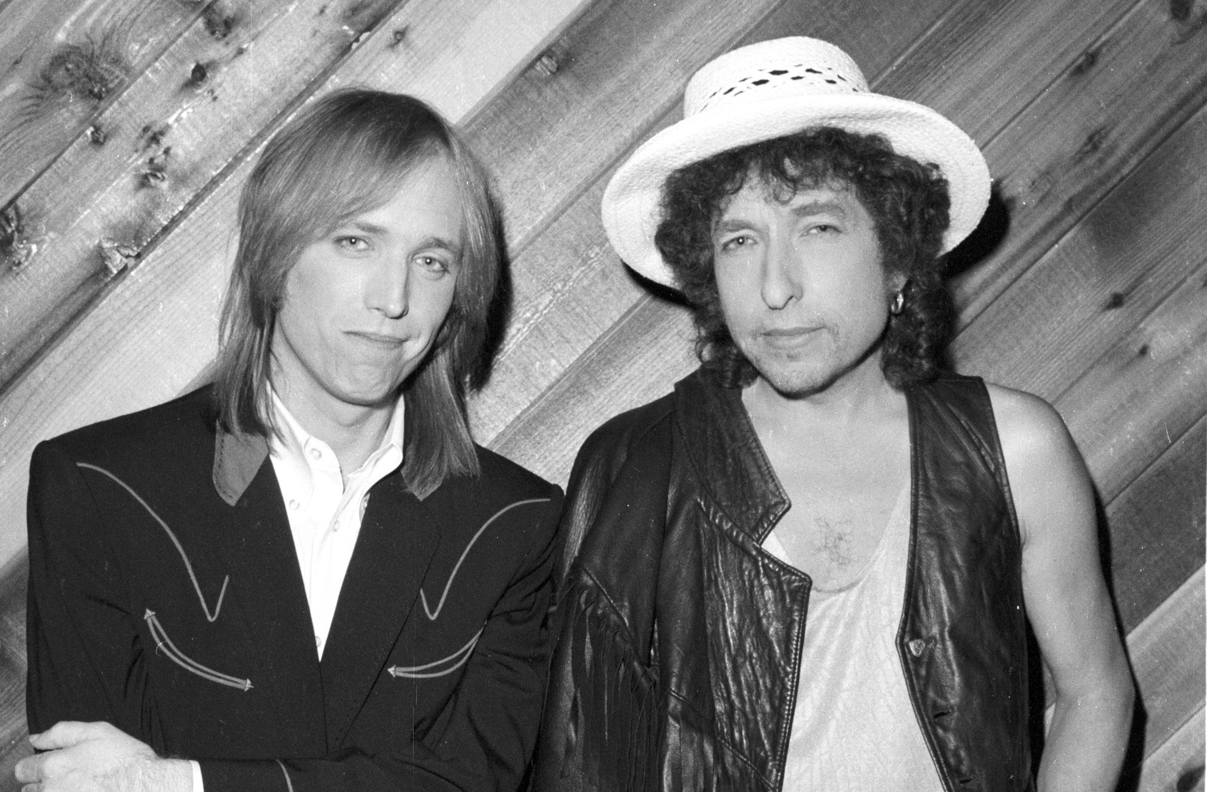 A black and white photo of Tom Petty and Bob Dylan standing in front of a wood paneled wall.