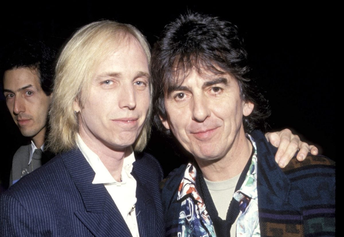 Tom Petty stands with his arm around George Harrison's shoulders against a black background.