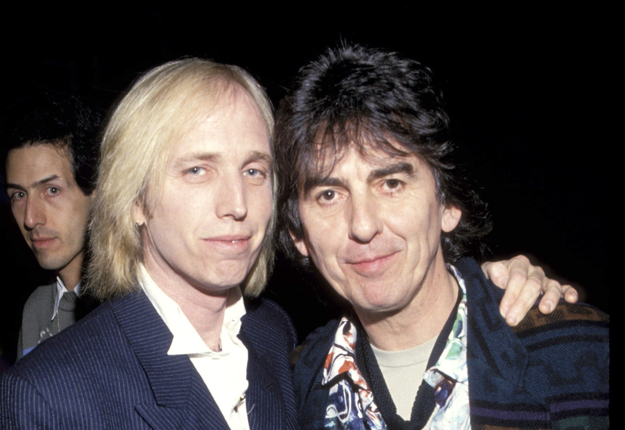 Tom Petty stands with his arm around George Harrison's shoulders.
