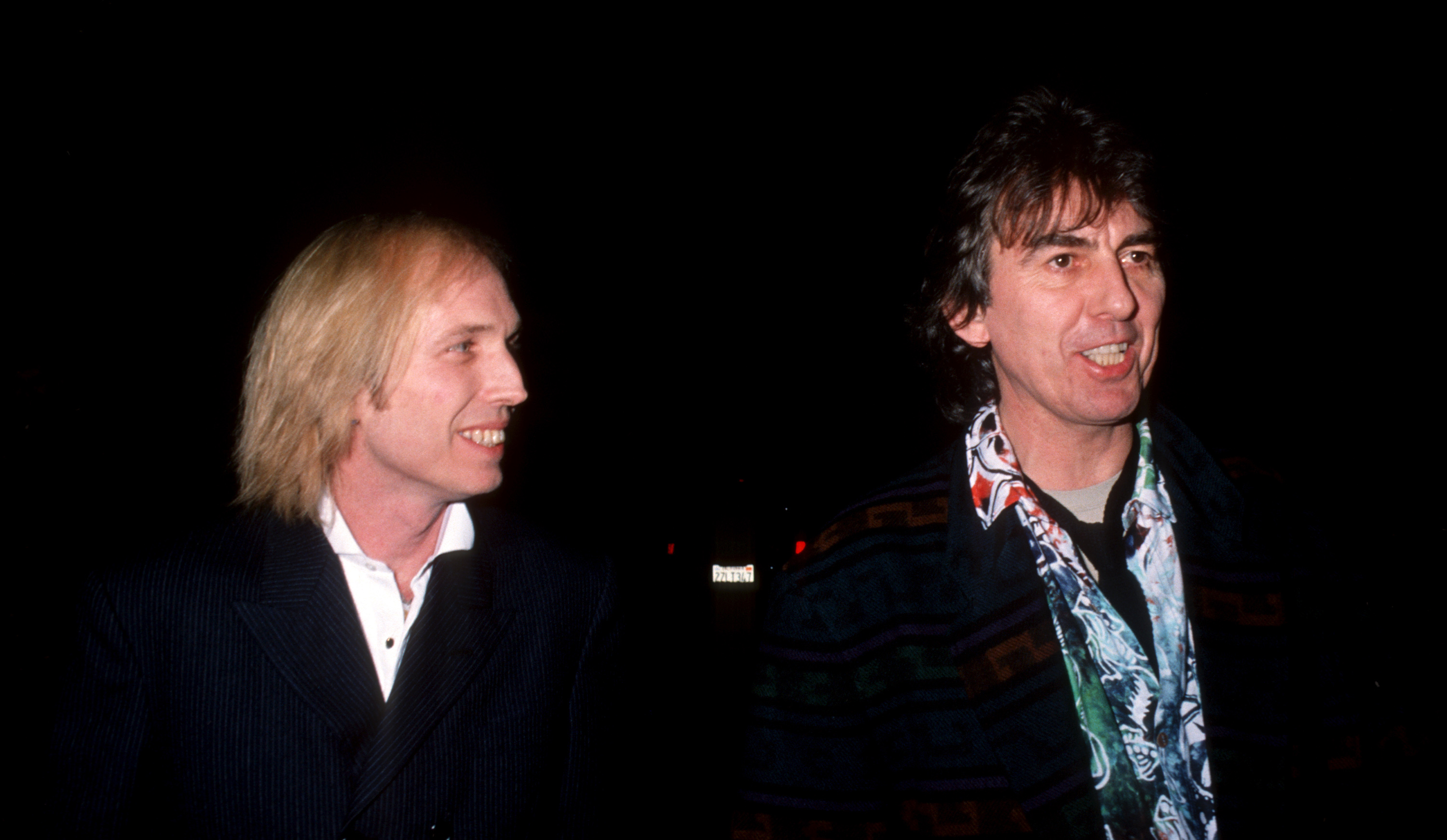 Tom Petty and George Harrison wear black jackets and walk together outside.