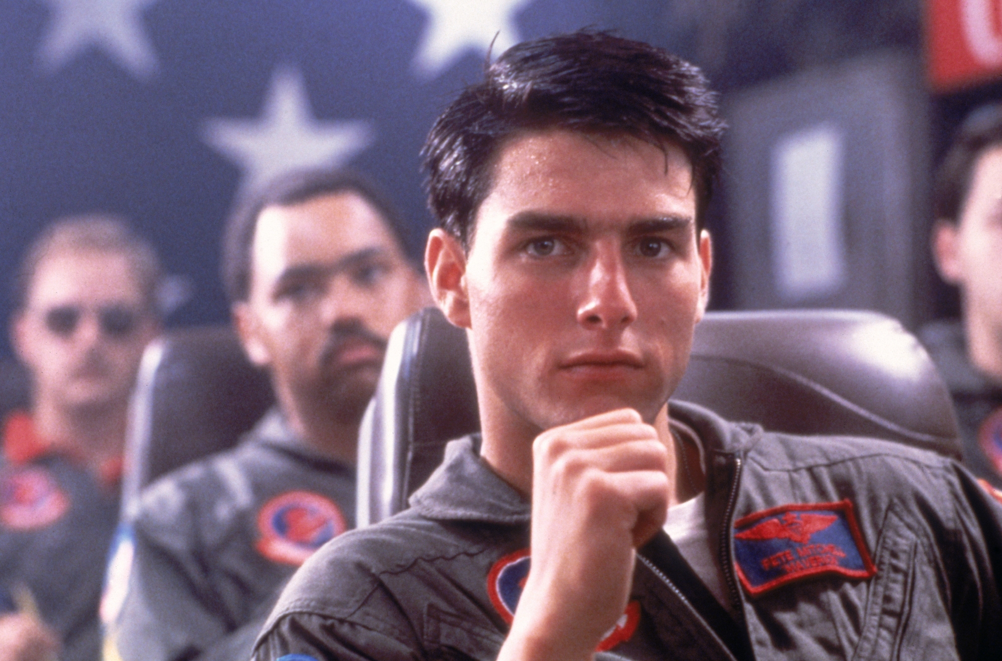 'Top Gun' Tom Cruise as Maverick holding his fist under his chin in front of the American flag and other uniformed actors