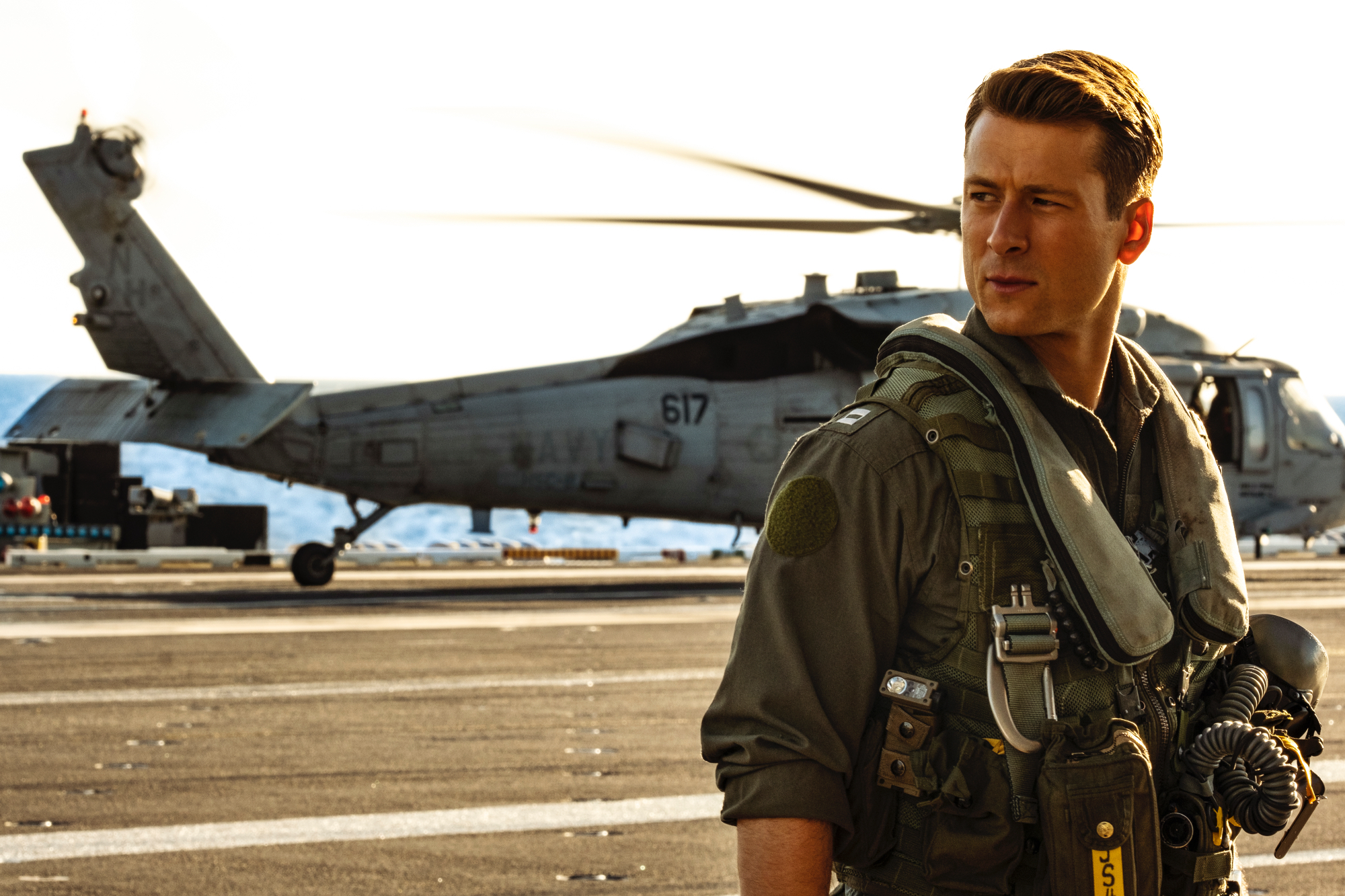 'Top Gun: Maverick' Glen Powell as Hangman wearing his pilot uniform and looking to the side with a helicopter in the background