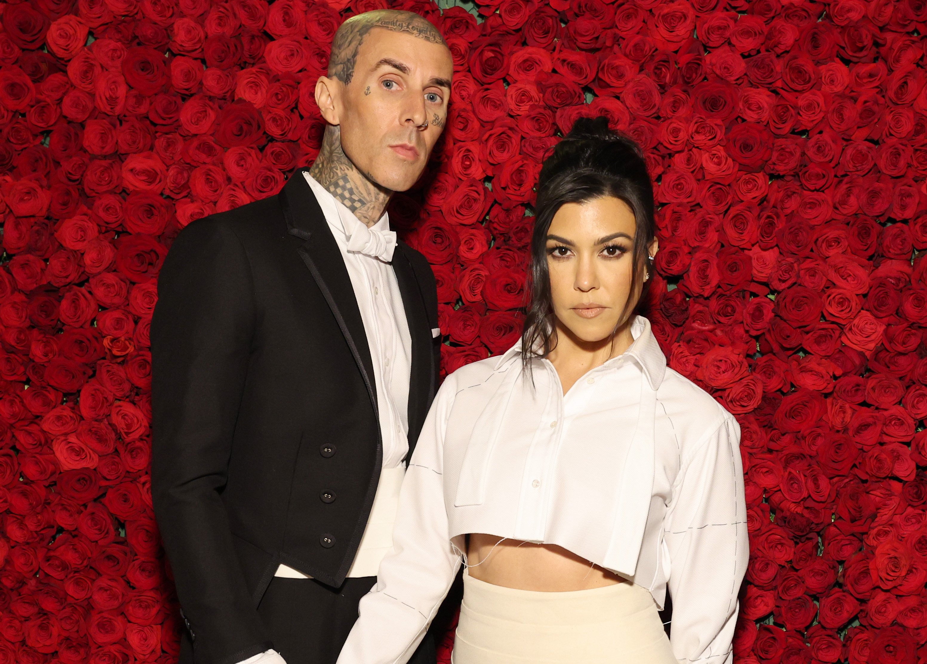 Travis Barker and Kourtney Kardashian pose for a photo in front of a wall of red flowers.