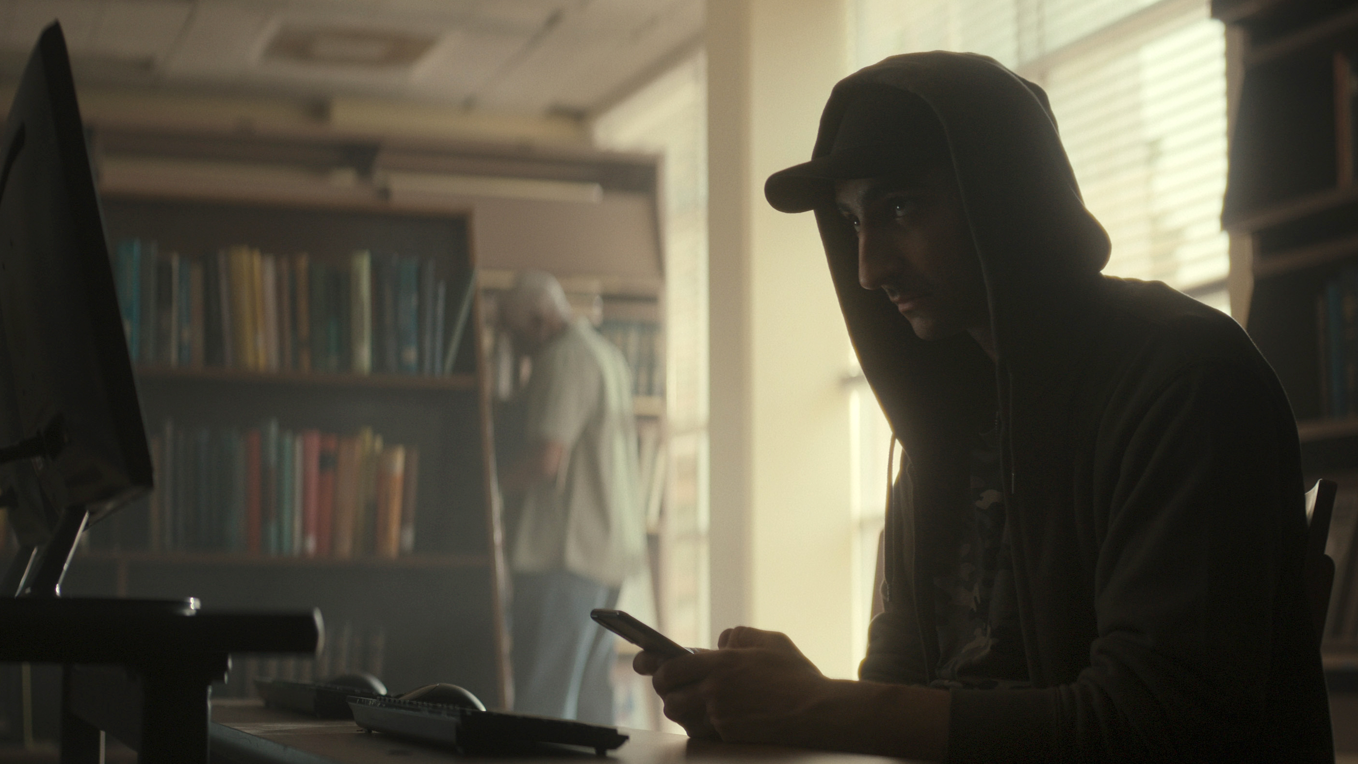 Man wearing a hoodie sitting in a library on his cell phone; screen grab from 'Web of Make Believe: Death, Lies, and the Internet'