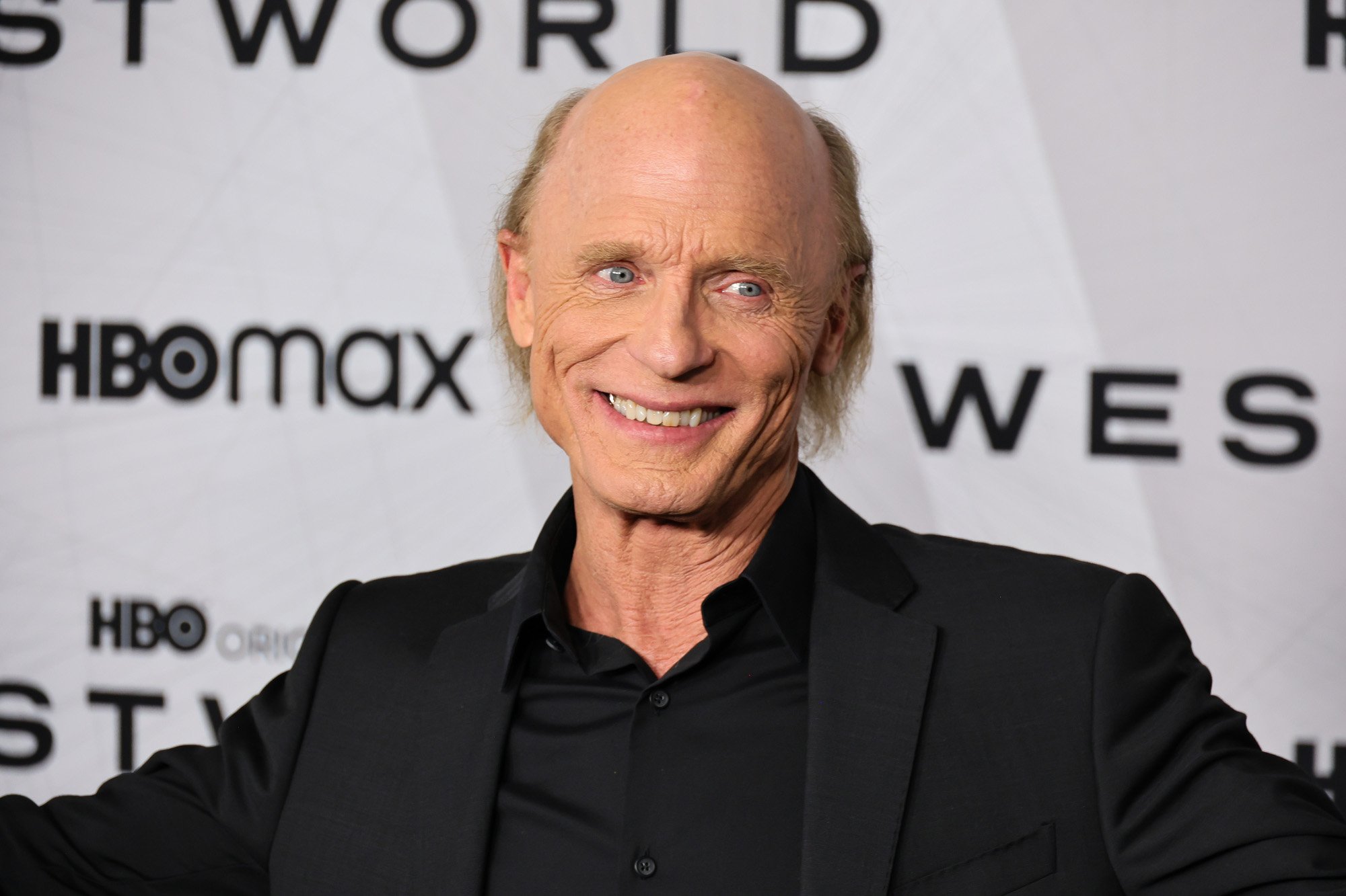 'Westworld' Season 4 star Ed Harris. He's wearing a black suit, holding his arms out, and smiling.
