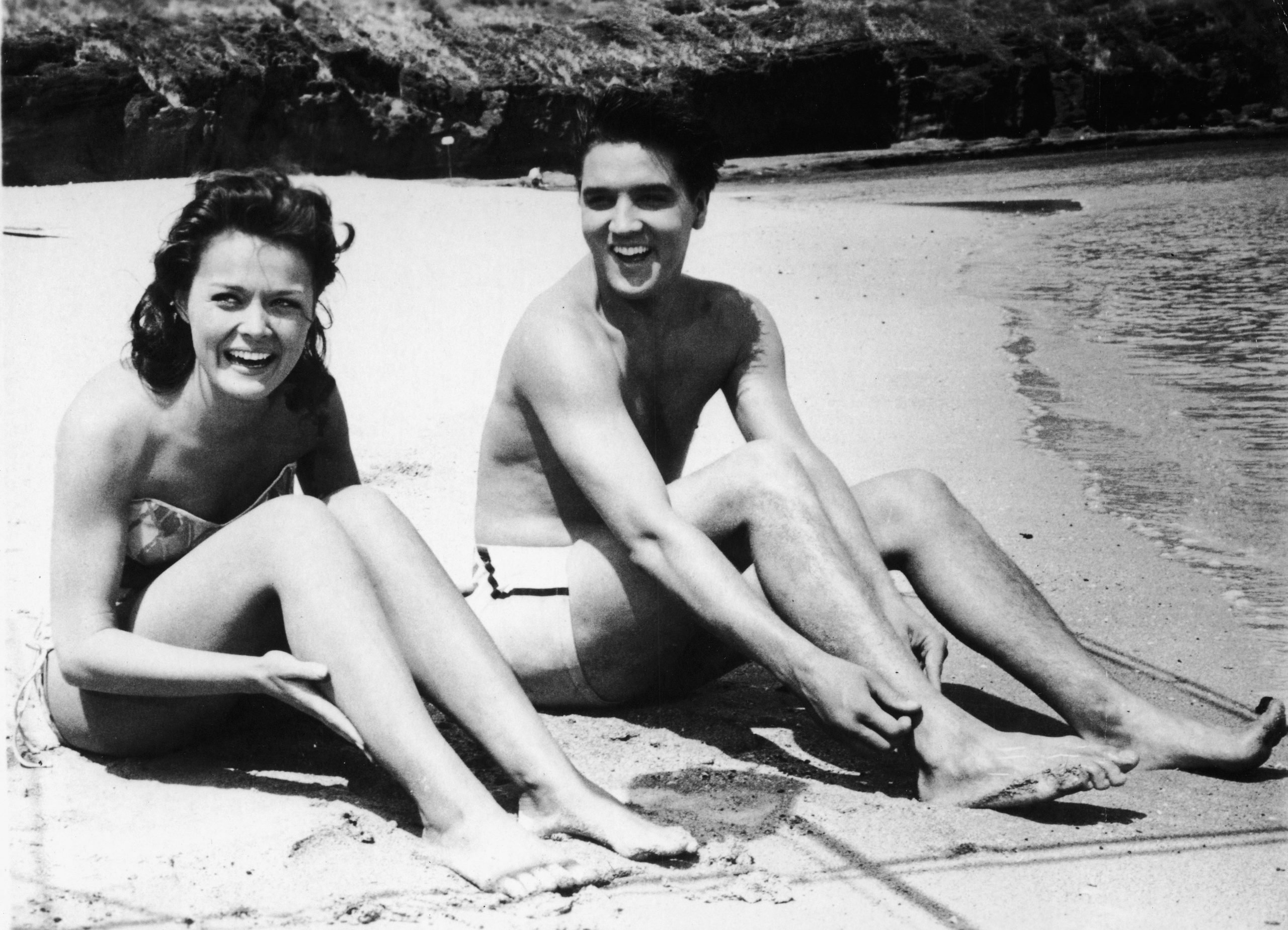 Joan Blackman and Elvis Presley sitting on a beach in a black-and-white image